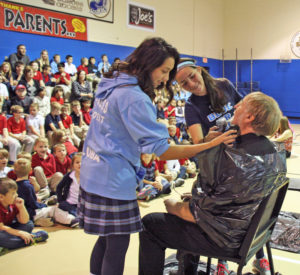 Anne Marie Amacher Sophia Gluba, left, and Sophia Kersten shave the beard off Deacon Chuck Metzger Nov. 30 at Lourdes Catholic School in Bettendorf. Deacon Metzger and teacher Alec Clark participated in No Shave November. 